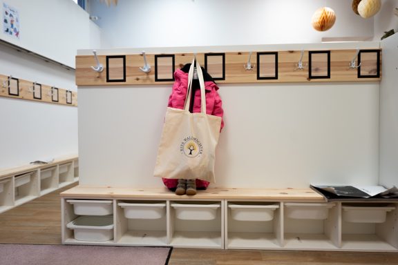 Rosa Jacke und Tasche hängen an einer Holzleiste vor einem hellen, minimalistischen Raum.